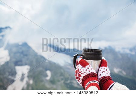 Closeup photo of thermos mug with tea in traveler's hand over out of focus mountains view with snow, tourizm in cold season