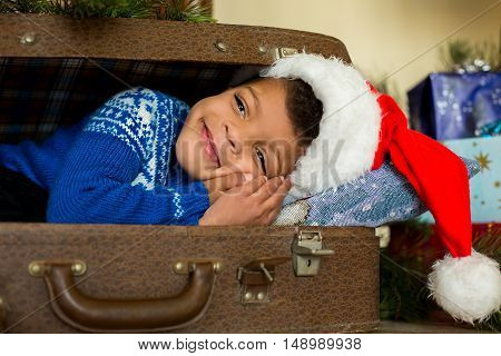 Little Santa in blue sweater. Darkskinned child near Christmas presents. Feeling warm and comfortable. So young and so happy.