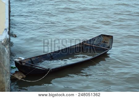 A Small Boat Is Tied To A Pier. It Is Half-filled With Water, And Covered With Rust. The Water Is Ca