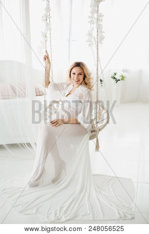 Beautiful Blonde Pregnant Woman Wearing In White Long Dress Sitting At Swing With Flowers And Lookin