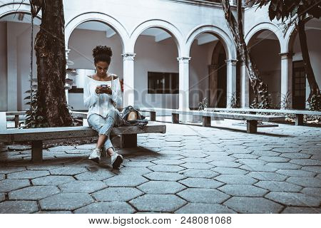 Wide-angle View Of A Pensive Cute African-american Female Student Who Is Sitting On The Concrete Ben