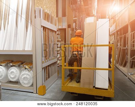 Worker In Helmet With Safety Belt On Forklift With Goods In Big Building Supermarket. Storage At War