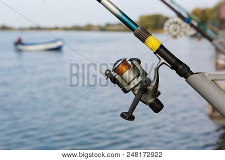 Fishing Rod With Spool Spinning Stands On The Bracket, Close-up, Against The Background Of Water, Fi