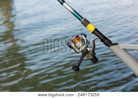 Fishing Rod With Spool Spinning Stands On The Bracket, Close-up, Against The Background Of Water