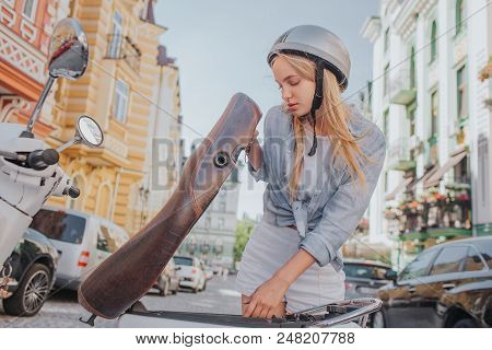Smart And Good-looking Girl Is Looking Under Motorcycle Seat. She Is Looking For Something. Girl Is 