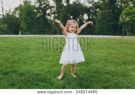 Smiling Little Cute Child Baby Girl In Light Dress Walking And Spreading Hands, Have Fun On Green Gr