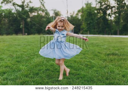 Pretty Little Cute Child Baby Girl In Denim Dress Circling Around And Have Fun On Green Grass Lawn I