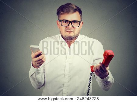 Young Man Holding Old-fashioned Red Phone And Modern Smarpthone Looking At Camera In Puzzlement.