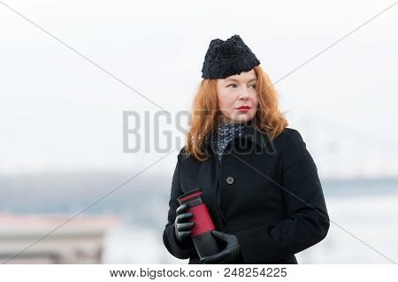 Ginger Woman In Black Coat And Hat With Red Lips. Woman Holding Thermos. Red Hair Lady With Cup In H