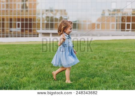 Smiling Little Cute Child Baby Girl In Denim Dress Walking And Running, Have Fun On Green Grass Lawn