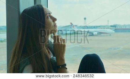 Young Sad Woman Is Sitting On The Window Sill At Airport, Crying And Wipes Tears Away With Airplane 