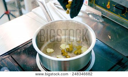 Hand Of Chef In Gloves Puts A Pea In Sauce Pan With Cooking Chopped Garlic. Food Is Steaming And His