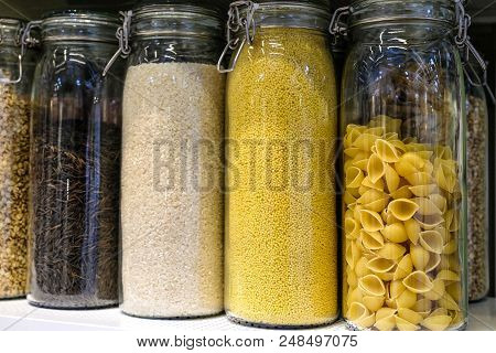 Various Raw Cereals, Grains, Beans And Pasta For Healthy Cooking In Glass Jars On Kitchen Shelf, Whi