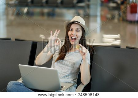 Young Surprised Traveler Tourist Woman In Hat Sitting, Working On Laptop, Holding Bitcoin, Showing O