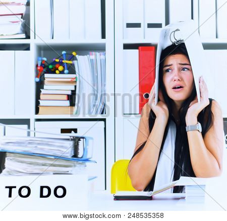 Funny Overworked Girl With A Folder On Her Head Sitting In The Workplace Cluttered With Folders. Rea