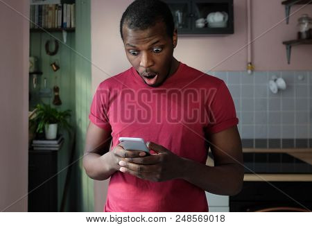 Closeup Portrait Of Handsome African Man Standing In His Kitchen Holding Mobile Phone In Both Hands 