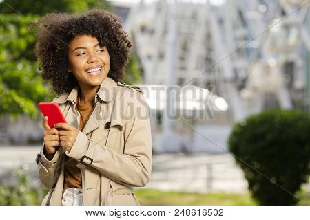 Mood-boosting Chat. Pleasant Curly Girl In A Trench Coat Smiling Broadly While Using Her Phone And T