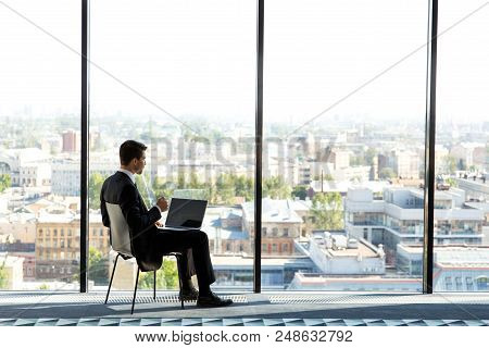 Business Man Sitting In Modern Office With Panoramic Windows, Typing On His Laptop And Srinking Coff