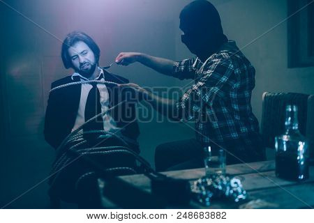 Man In Mask Is Holding Big Spoon Close To Victim's Face. Businessman Is Sitting With His Body Tied T