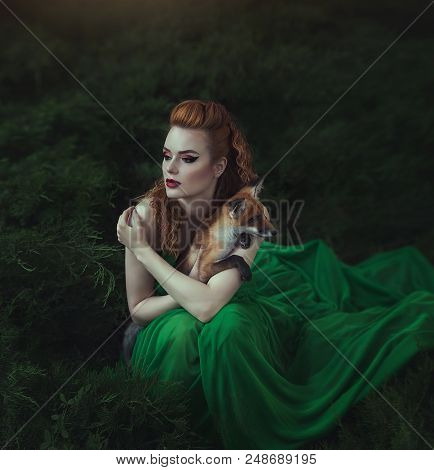 A Red-haired Girl In A Green Dress Is Sitting With A Fox In A Fairy Forest. Beautiful Young Woman Wi