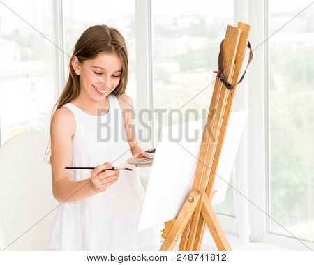 Pretty girl with brush in hand. Creative teen girl paitning a picture on easel. Girl practising drawing in art school