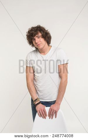 Handsome Young Man With Curly Hair Standing In White T-shirt Behind A Chair.