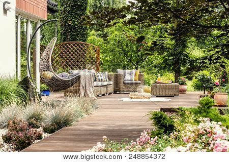 Hanging Chair And Pouf On Wooden Veranda In The Middle Of Garden With Flowers. Real Photo