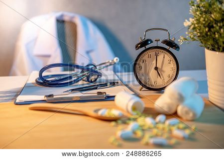 Stethoscope, Clipboard, Pen And Table Clock On Dector Desk. Blurred Pills Spilling Out Of Pill Bottl