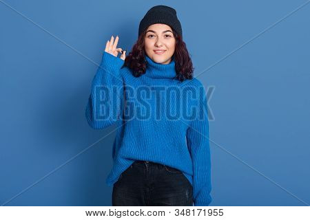 Indoor Picture Of Positive Cute Pleasant Brunette Making Gesture, Showing Sign Of Okay, Looking Dire