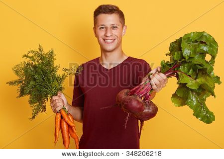 Inddor Shot Of Man In Dresses Burgundy T Shirt Holding Bunch Of Fresh Organic Young Carrots And Bunc
