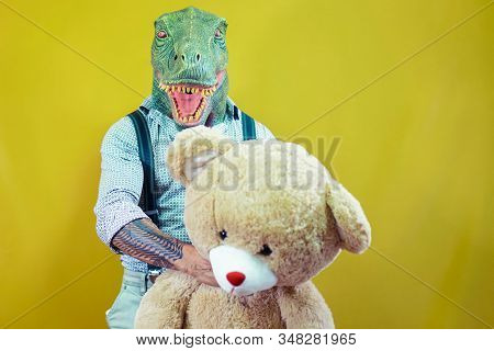 Senior Man Wearing T-rex Dinosaur Mask With Teddy Bear. Crazy Man Having Fun Celebrating Carnival An