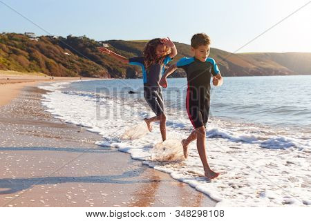 Two Children Wearing Wetsuits Running Through Waves On Summer Beach Vacation