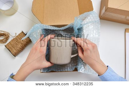 delivery, mail service, people and shipment concept - close up of female hands packing mug into parcel box and it wrapping into protective bubble wrap at post office