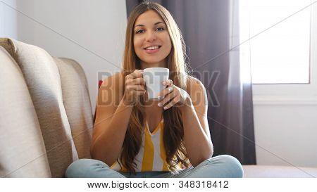 Happy Student Girl Sitting On Sofa With Crossed Legs Holding A Mug Of Tea Or Cappuccino At Home In T