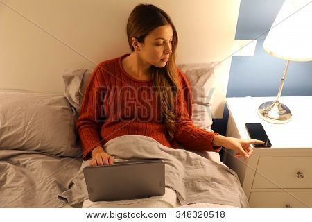 Student Girl Working On Laptop On Bed Distracted By Phone At The Night Time