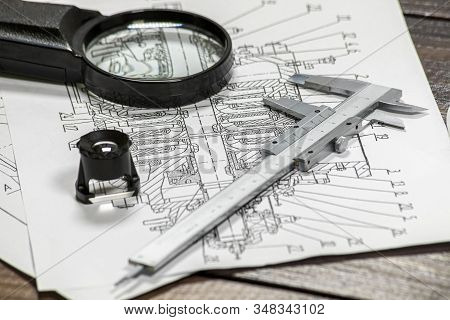 Some Pump Blueprints On A Wooden Desk With Two Magnithing Glasses And A Calipers.