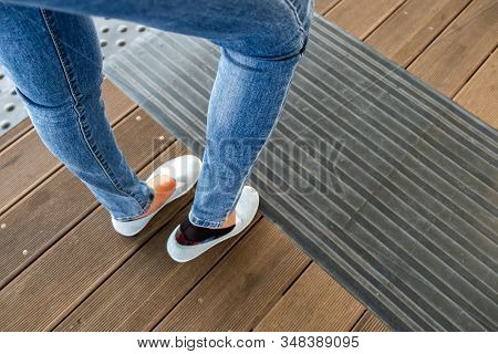 Female Legs In Denim Jeans Pants And Light Summer Sneakers Standing On Wooden Floow.