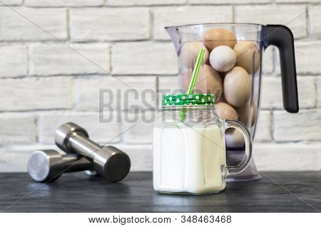 Protein Shake In A Mug Against The Background Of A Blender Bowl With Chicken Eggs And Dumbbells Agai
