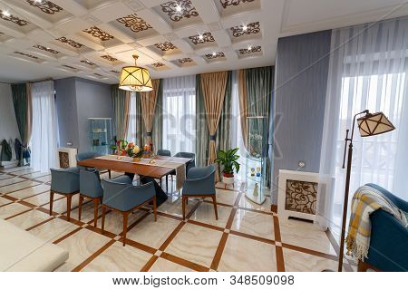 Living room of a large country house with a large table for eight seats. Interior with beautiful curtains. Chandelier lights on. Square ceiling.