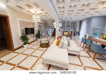 Living room of a large country house with a large table for eight seats. A sofa in the middle of the room with a TV hanging on the wall. Chandelier lights on. View of the kitchen and stairs.