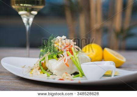 White Fish Seabass Fillet With Vegetables And White Wine On A Wooden Table In A Restaurant With Deco