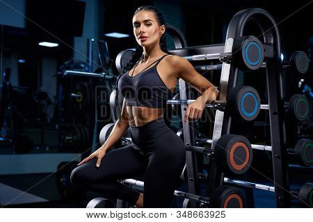 Side Close Up Of Female Bodybuilder With Long Braids Posing Near Stand With Sports Equipment. Srtong