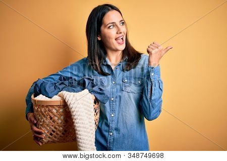 Young brunette woman with blue eyes doing domestic chores holding laundry basket pointing and showing with thumb up to the side with happy face smiling
