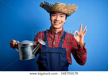 Young handsome chinese farmer man wearing apron and straw hat holding watering can doing ok sign with fingers, excellent symbol