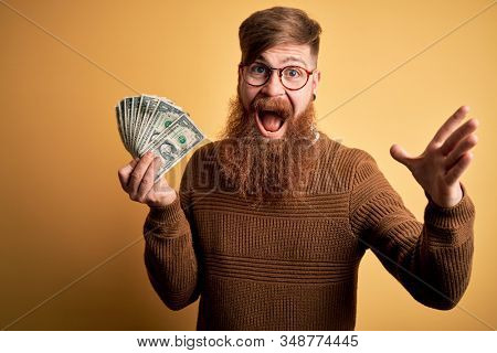 Redhead Irish man with beard holding a bunch of 1 dolla banknotes over yellow background very happy and excited, winner expression celebrating victory screaming with big smile and raised hands