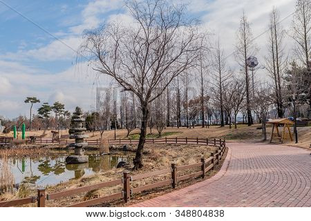 Leafless Tree In Front Of Stone Tower In Pond In Urban Public Park On Partly Cloudy Winter Day.
