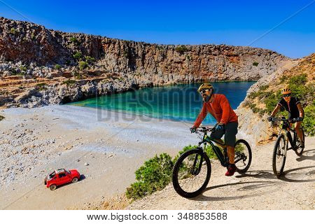 Cycling woman and man on electric bikes to wild beach with turquoise water and off-road  access . Couple cycling MTB enduro trail track. Outdoor sport activity.