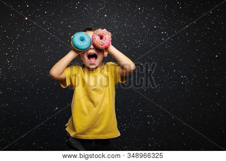 Happy cute boy is having fun played with donuts on black background wall. Bright photo of a boy. Colored donuts