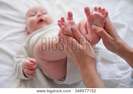 Baby Feet In Mother Hands. Young Caucasian Woman Makes Massage For Happy Infant Baby On White Bed At