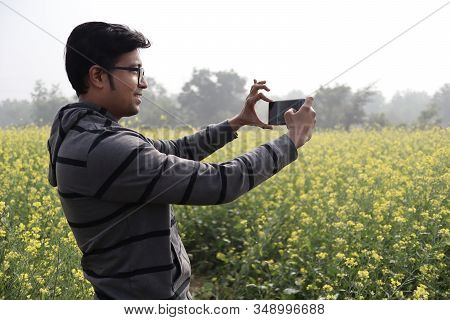 A Man In Hoodie And Spectacles Clicking Nature Photo With His Smartphone In A Mustard Field With Yel
